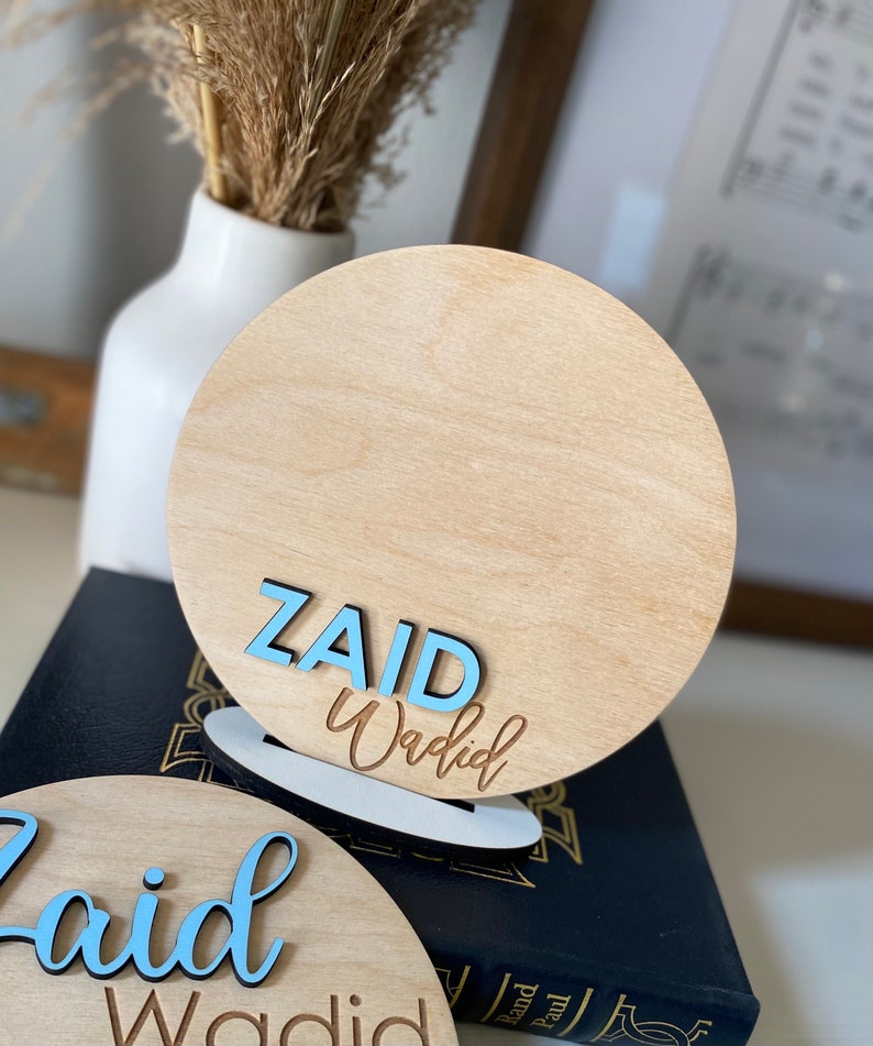 May include: Two wooden round signs with the name "Zaid Waqdid" written in blue and brown lettering. The signs are on a black surface.