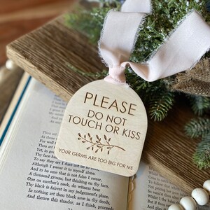 May include: An arched wooden sign with a light wood grain finish, featuring the engraved text "PLEASE DO NOT TOUCH OR KISS" and "YOUR GERMS ARE TOO BIG FOR ME" with a small leaf illustration. A pale pink frayed ribbon is tied through the top hole.