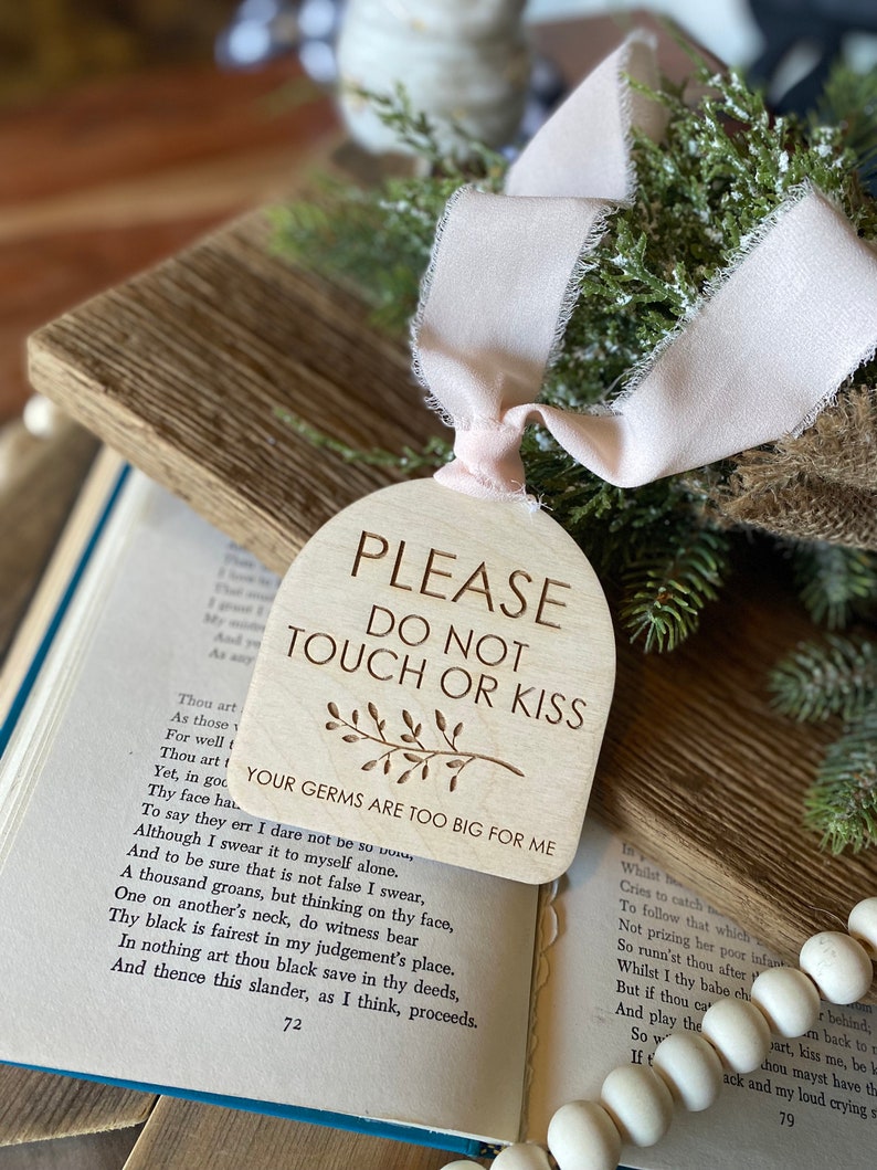 May include: An arched wooden sign with a light wood grain finish, featuring the engraved text "PLEASE DO NOT TOUCH OR KISS" and "YOUR GERMS ARE TOO BIG FOR ME" with a small leaf illustration. A pale pink frayed ribbon is tied through the top hole.