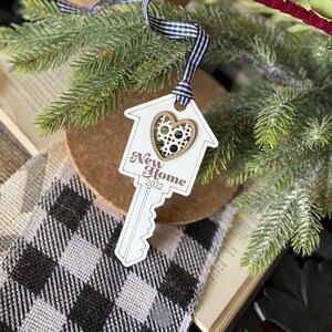 May include: White wooden key-shaped ornament with a house cutout and a heart in the center. The ornament says "New Home 2022" and is tied with a black and white ribbon.