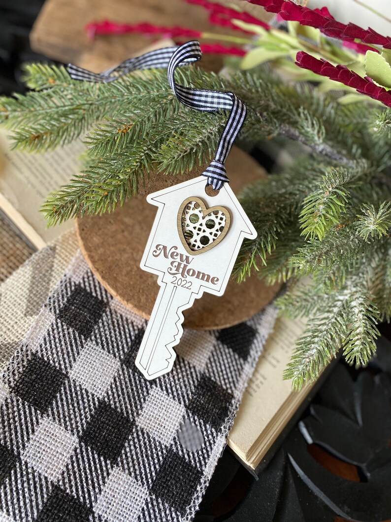 May include: White wooden key-shaped ornament with a house cutout and a heart in the center. The ornament says "New Home 2022" and is tied with a black and white ribbon.