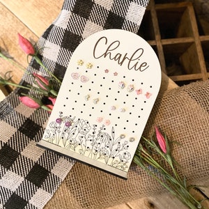 May include: A white wooden earring holder with a floral design and the name "Charlie" written in cursive. The holder has a rounded top and a flat base. It is decorated with small black dots and features a variety of colorful earrings.