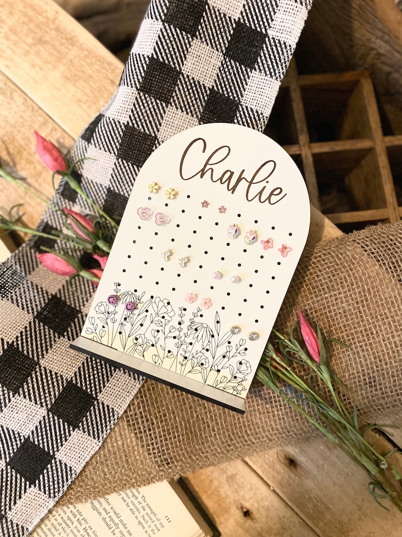 May include: A white wooden earring holder with a floral design and the name "Charlie" written in cursive. The holder has a rounded top and a flat base. It is decorated with small black dots and features a variety of colorful earrings.