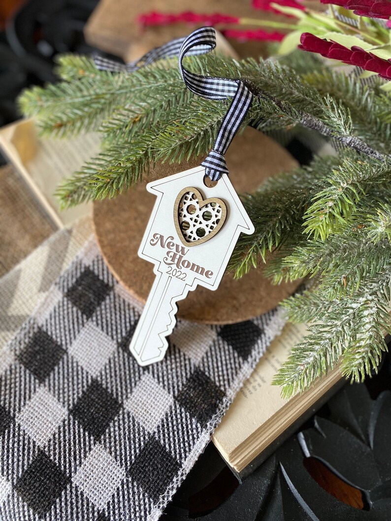 May include: A white wooden key-shaped ornament with a house cutout and a heart-shaped cutout in the center. The ornament is decorated with the words "New Home 2022".