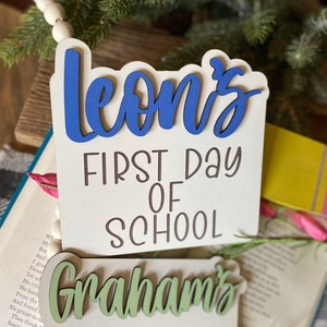 May include: Two wooden signs with the text "Leon's First Day of School" and "Graham's First Day of" in brown lettering. The name "Leon's" is painted in blue and "Graham's" is painted in green.