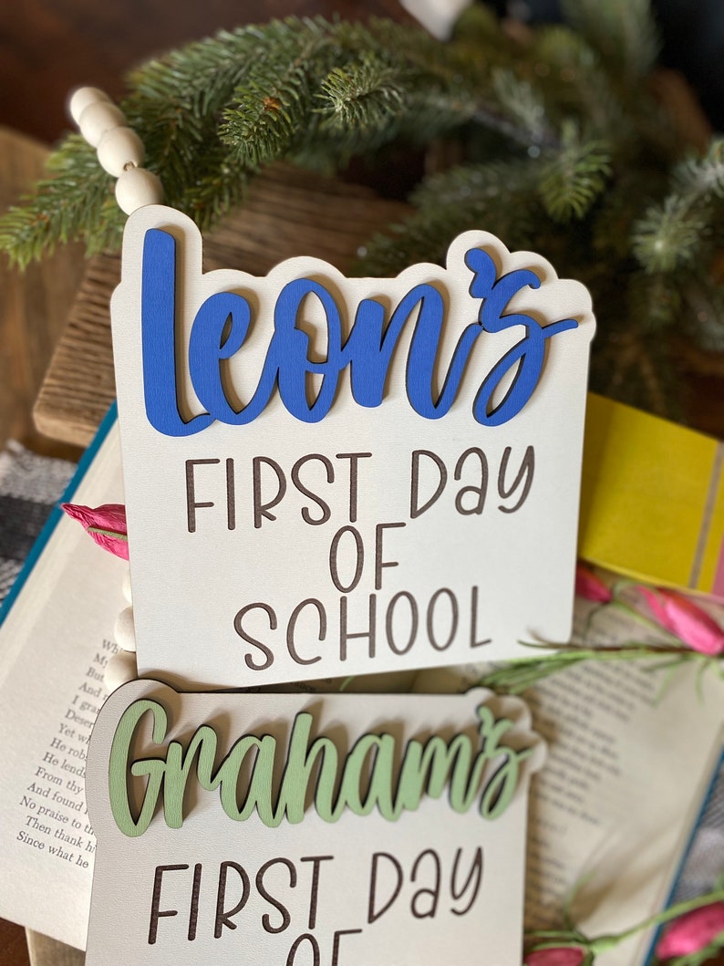 May include: Two wooden signs with the text "Leon's First Day of School" and "Graham's First Day of" in brown lettering. The name "Leon's" is painted in blue and "Graham's" is painted in green.