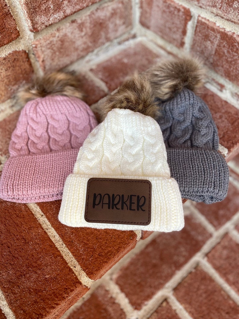 Op de afbeelding: Drie gebreide mutsen met nep bont pompoenen. E&eacute;n muts is roze, &eacute;&eacute;n is wit met een bruin leren patch waarop "PARKER" staat, en &eacute;&eacute;n is grijs.