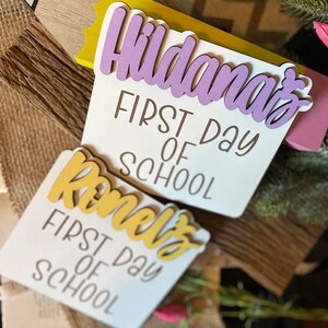 May include: Two white wooden signs with the text "First Day of School" and the names "Hildana's" and "Romelo's" in purple and yellow lettering.