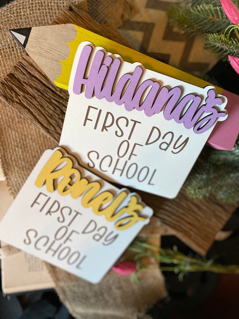 May include: Two white wooden signs with the text "First Day of School" and the names "Hildana's" and "Romelo's" in purple and yellow lettering.