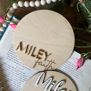 May include: Two wooden round signs with the names "Miley Faith" written in white and gold lettering. The signs are blank and ready to be personalized with a date and time.