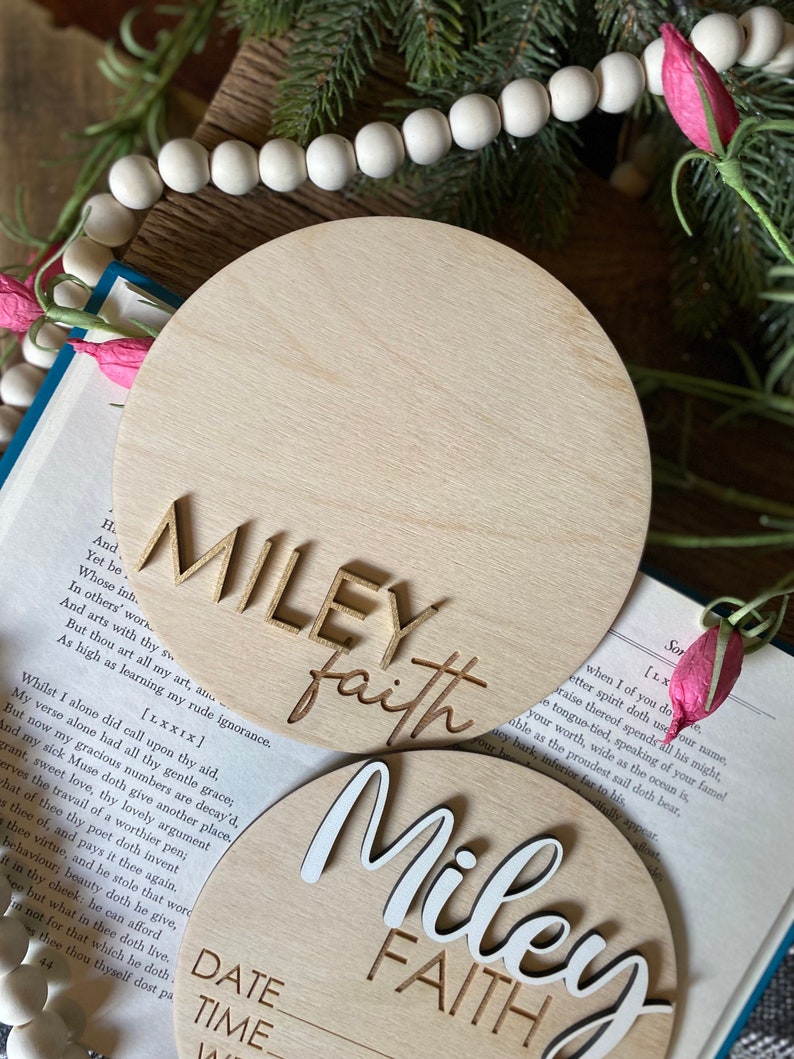 May include: Two wooden round signs with the names "Miley Faith" written in white and gold lettering. The signs are blank and ready to be personalized with a date and time.