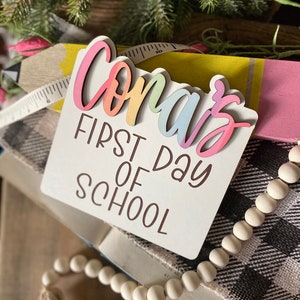 May include: A white wooden sign with the text "Conar's First Day of School" in brown lettering. The word "Conar's" is in rainbow colors and is layered on top of the sign.