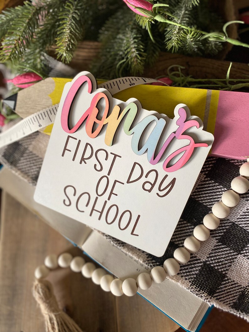 May include: A white wooden sign with the text "Conar's First Day of School" in brown lettering. The word "Conar's" is in rainbow colors and is layered on top of the sign.