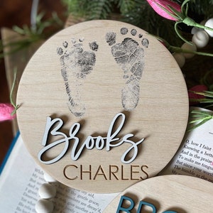 May include: A round wooden sign with a light brown background. The sign features two black baby footprints and the names "Brooks Charles" in white and brown lettering.