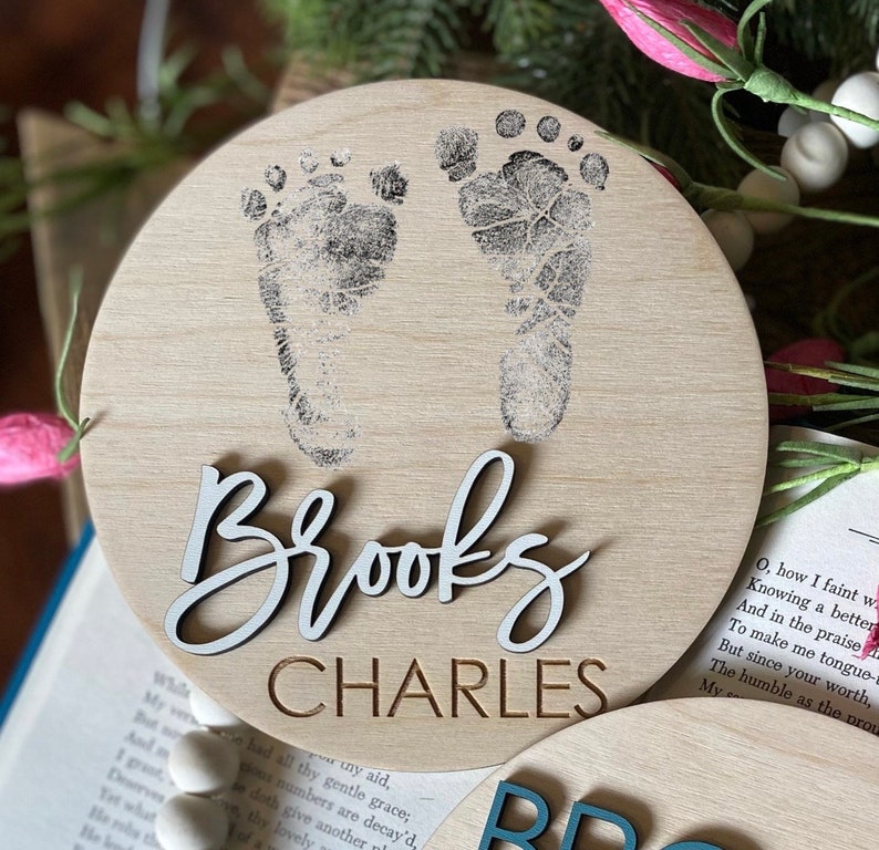 May include: A round wooden sign with a light brown background. The sign features two black baby footprints and the names "Brooks Charles" in white and brown lettering.