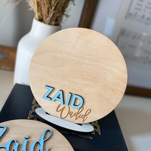 May include: Two wooden round signs with the name "Zaid Waqdid" written in blue and brown lettering. The signs are on a black surface.