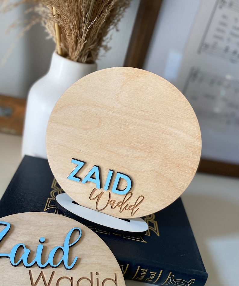 May include: Two wooden round signs with the name "Zaid Waqdid" written in blue and brown lettering. The signs are on a black surface.