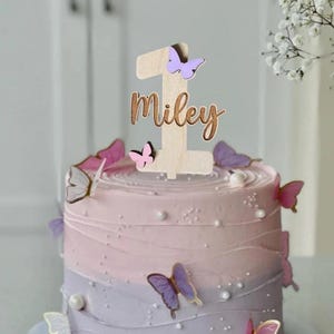 May include: A wooden cake topper with the number one and the name Miley, decorated with purple and pink butterflies. The topper is on a two-toned purple and pink cake.