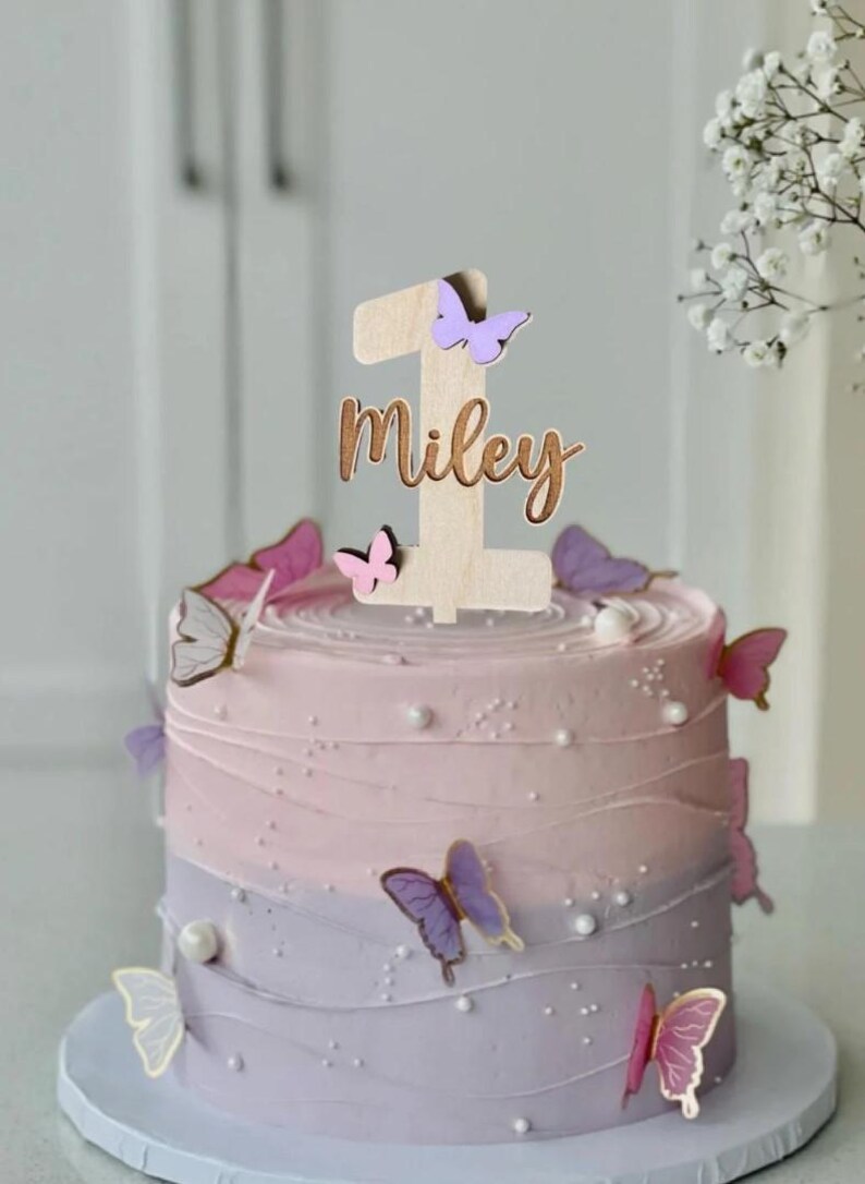 May include: A wooden cake topper with the number one and the name Miley, decorated with purple and pink butterflies. The topper is on a two-toned purple and pink cake.