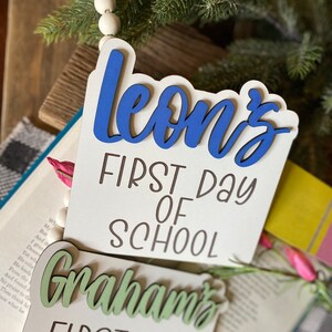 May include: Two wooden signs with the text "Leon's First Day of School" and "Graham's First Day of" in black lettering. The name "Leon's" is in blue and the name "Graham's" is in green.
