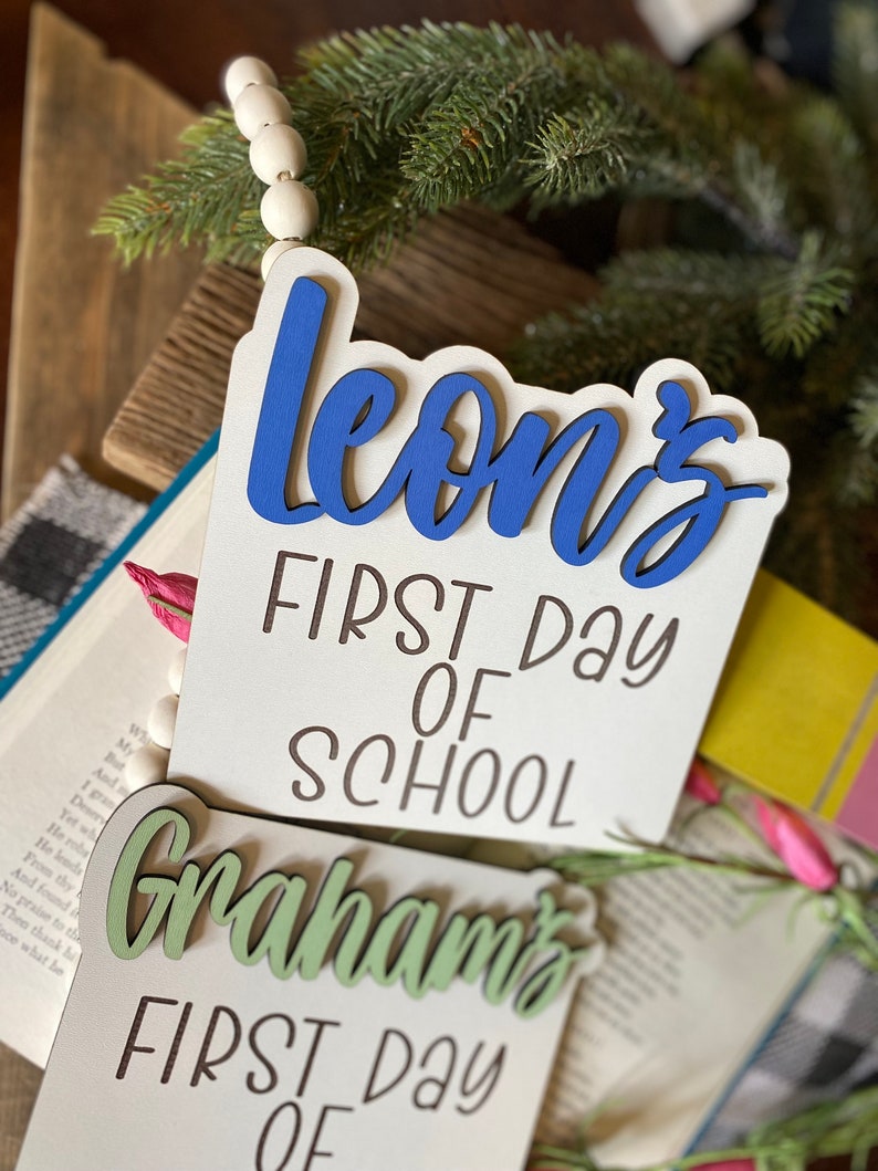May include: Two wooden signs with the text "Leon's First Day of School" and "Graham's First Day of" in black lettering. The name "Leon's" is in blue and the name "Graham's" is in green.