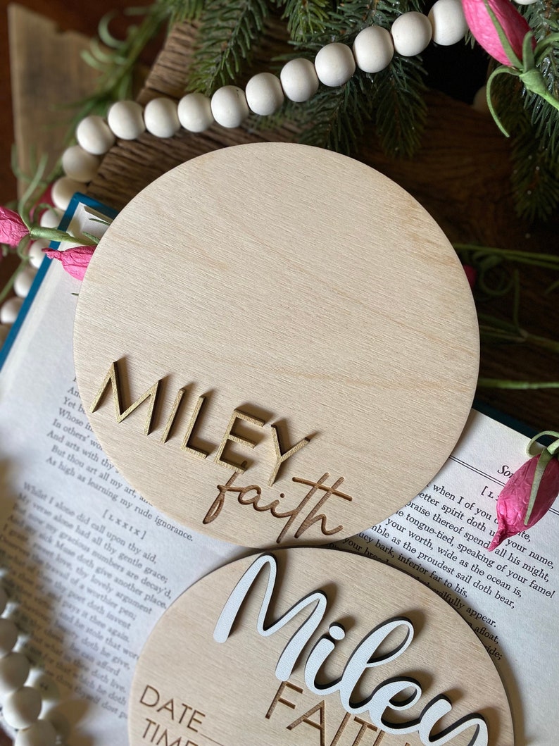 May include: Two wooden round plaques with the names "MILEY faith" and "MILEY FAITH" in gold and white lettering. The plaques are on a book with a white and pink flower.