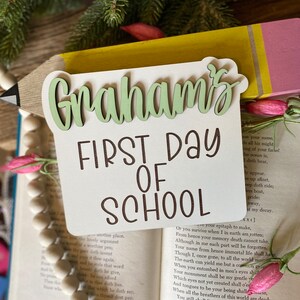 May include: A wooden sign with the text "Graham's First Day of School" in brown lettering. The sign is shaped like a pencil with a yellow body and a pink eraser. The sign is resting on a book with a pink flower on the cover.