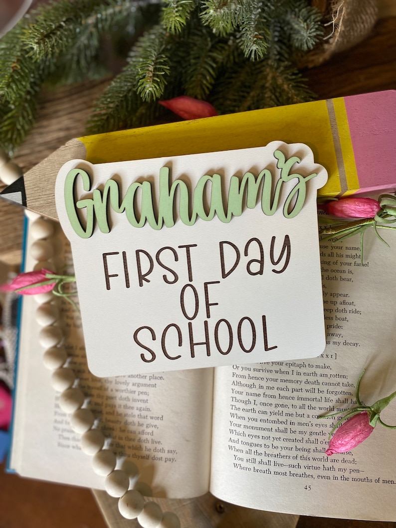 May include: A wooden sign with the text "Graham's First Day of School" in brown lettering. The sign is shaped like a pencil with a yellow body and a pink eraser. The sign is resting on a book with a pink flower on the cover.