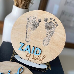 May include: A wooden round sign with a light brown background. The sign has two black baby footprints and the name "ZAID Wadid" written in blue letters.