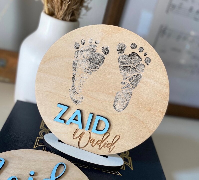May include: A wooden round sign with a light brown background. The sign has two black baby footprints and the name "ZAID Wadid" written in blue letters.