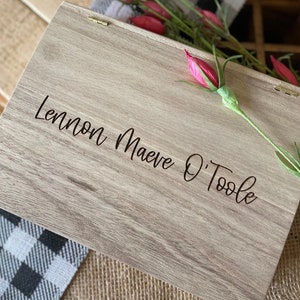 Puede incluir: Una caja de madera con tapa con bisagras, grabada con el nombre "Lennon Maeve O'Toole". La caja es de color marr&oacute;n claro con un patr&oacute;n de veta de madera. La caja est&aacute; sobre una tela a cuadros en blanco y negro.