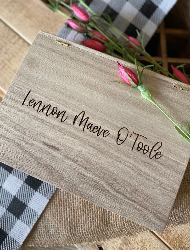 Puede incluir: Una caja de madera con tapa con bisagras, grabada con el nombre "Lennon Maeve O'Toole". La caja es de color marr&oacute;n claro con un patr&oacute;n de veta de madera. La caja est&aacute; sobre una tela a cuadros en blanco y negro.