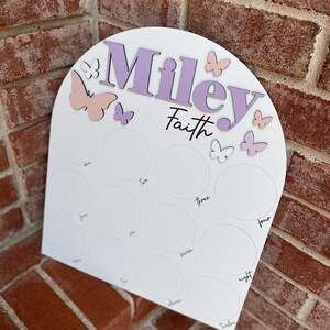 May include: A white wooden arch with the names "Miley Faith" written in purple and pink letters. The arch has 12 circles with numbers 1 through 12 written inside each circle. There are several pink and purple butterflies on the arch.