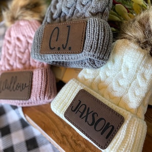 Op de afbeelding: Drie gebreide mutsen met nep bont pompoenen en gepersonaliseerde leren patches. De mutsen zijn roze, grijs en wit. De patches lezen "Willow", "C.J." en "Jaxson".