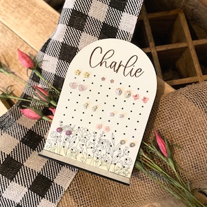 May include: White wooden earring holder with a floral design and black polka dots. The holder has a cutout arch at the top with the name "Charlie" written in cursive.