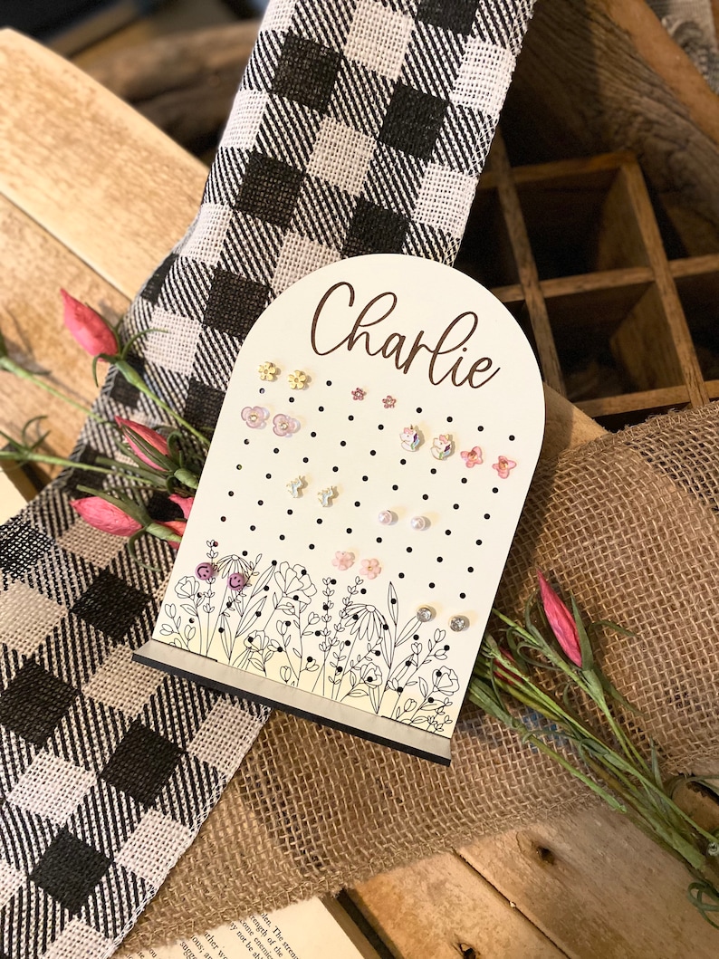 May include: White wooden earring holder with a floral design and black polka dots. The holder has a cutout arch at the top with the name "Charlie" written in cursive.