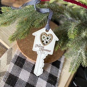 May include: White wooden key-shaped ornament with a house cutout and a heart in the center. The ornament has the text "New Home 2022" printed on it. The ornament is hanging from a black and white gingham ribbon.