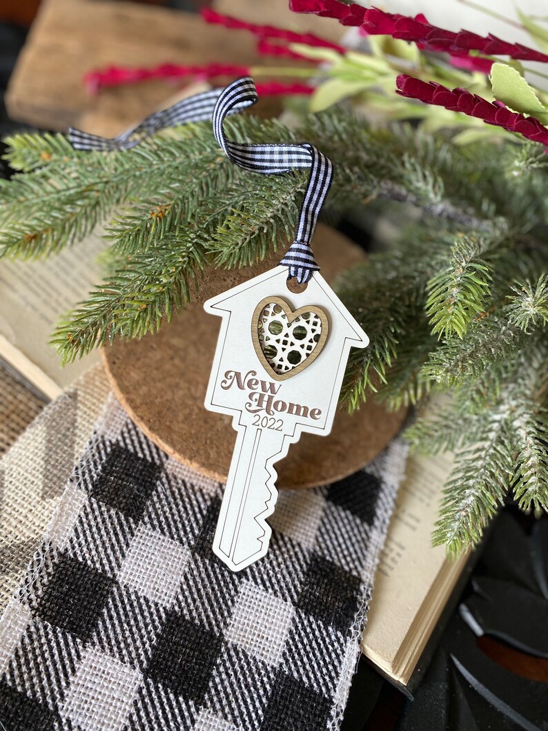 May include: White wooden key-shaped ornament with a house cutout and a heart in the center. The ornament has the text "New Home 2022" printed on it. The ornament is hanging from a black and white gingham ribbon.