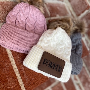 Op de afbeelding: Drie gebreide mutsen met nep bont pompoenen. E&eacute;n muts is roze, &eacute;&eacute;n is wit met een bruin leren patch waarop "PARKER" staat, en &eacute;&eacute;n is grijs.