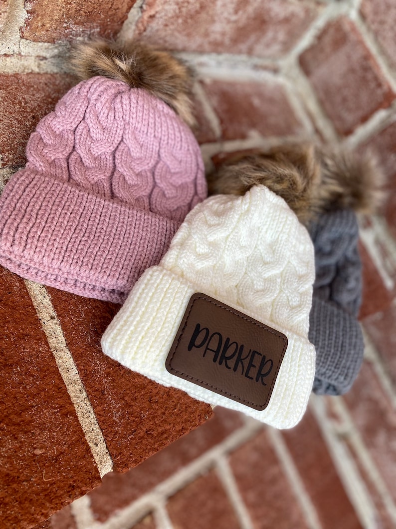 Op de afbeelding: Drie gebreide mutsen met nep bont pompoenen. E&eacute;n muts is roze, &eacute;&eacute;n is wit met een bruin leren patch waarop "PARKER" staat, en &eacute;&eacute;n is grijs.