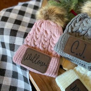 Op de afbeelding: Drie gebreide mutsen met nep bont pompoenen en gepersonaliseerde leren patches. De mutsen zijn roze, grijs en wit. De patches lezen "Willow", "C.J." en "Jackson".