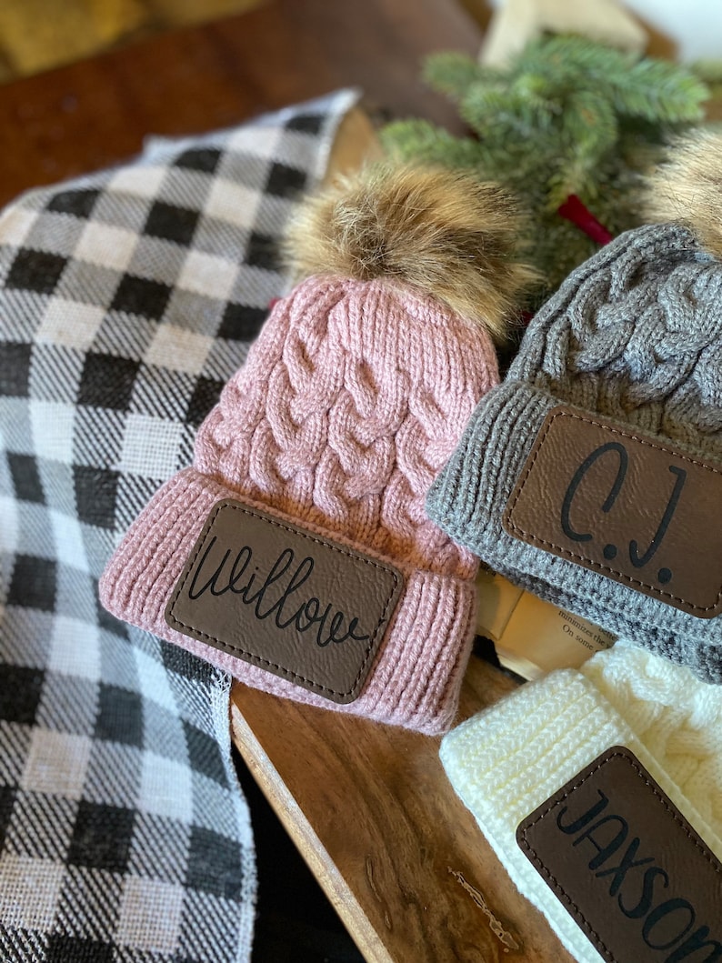 Op de afbeelding: Drie gebreide mutsen met nep bont pompoenen en gepersonaliseerde leren patches. De mutsen zijn roze, grijs en wit. De patches lezen "Willow", "C.J." en "Jackson".