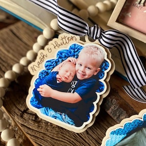 Op de afbeelding: Een houten ornament met een foto van twee broers. De oudere broer houdt de jongere broer vast. Het ornament is versierd met een gegolfde rand en de tekst "Nash & Hutton".