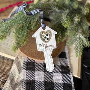 May include: White wooden ornament shaped like a house key with a heart cutout and the text "New Home 2022". The ornament is hanging from a black and white ribbon.