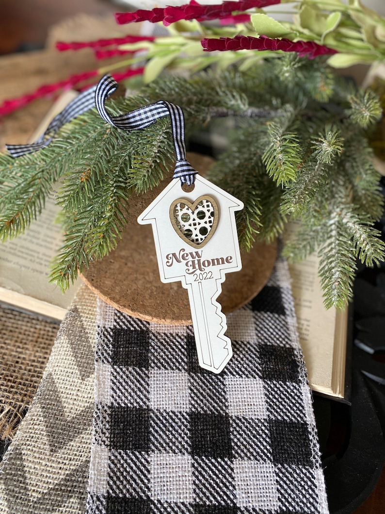May include: White wooden ornament shaped like a house key with a heart cutout and the text "New Home 2022". The ornament is hanging from a black and white ribbon.