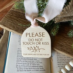 May include: An arch-shaped wooden sign with a light pink frayed ribbon. The text reads: PLEASE DO NOT TOUCH OR KISS, YOUR GERMS ARE TOO BIG FOR ME, with a small engraved branch illustration. The sign is positioned over an open book.