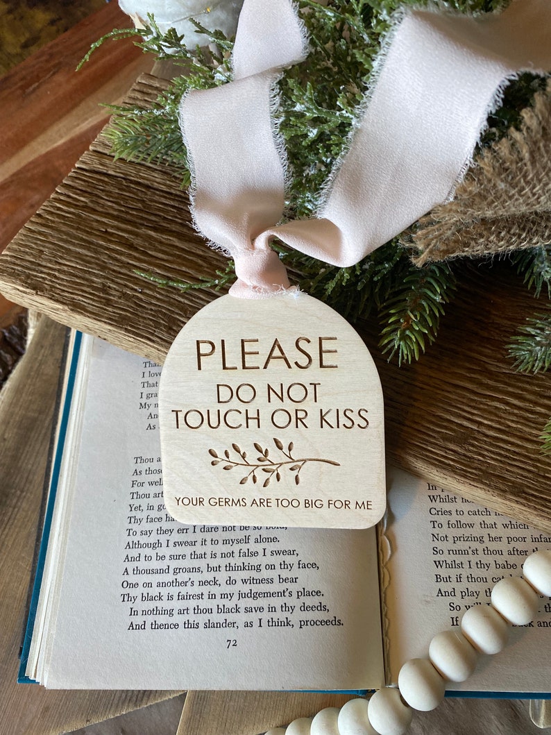 May include: An arch-shaped wooden sign with a light pink frayed ribbon. The text reads: PLEASE DO NOT TOUCH OR KISS, YOUR GERMS ARE TOO BIG FOR ME, with a small engraved branch illustration. The sign is positioned over an open book.