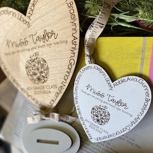 May include: Two wooden heart-shaped ornaments with the text "Miss Taylor" and "Thank you for putting your heart into teaching". Each ornament features a small apple design and the text "4th Grade Class 2023-2024" and "6th Grade Class 2023-2024". The ornaments are surrounded by a white surface and a string of white beads.
