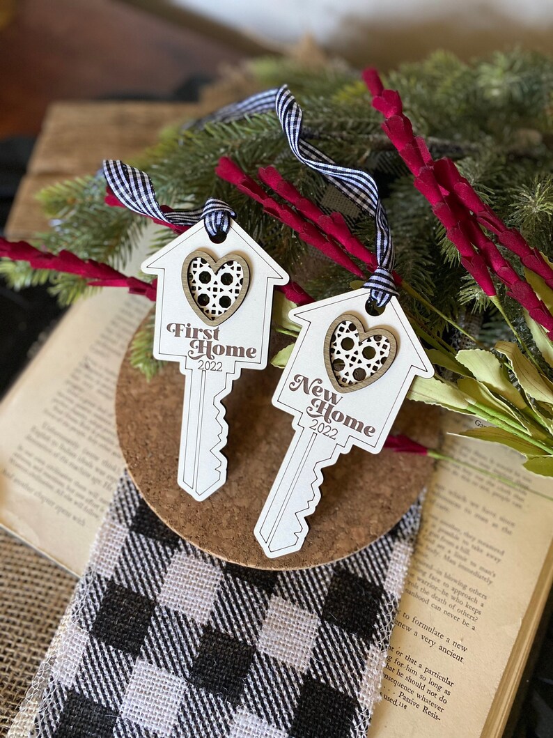 May include: Two wooden key-shaped ornaments with a house design and a heart in the center. The ornaments are white with brown accents and the text "First Home 2022" and "New Home 2022" is printed on them.