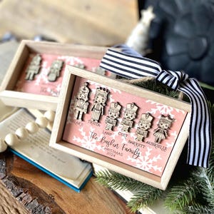 Könnte beinhalten: Ein rosa-weißes Ornament aus Holz mit einem rosa Hintergrund und weißen Schneeflocken. Das Ornament zeigt fünf Holz-Nussknackerfiguren mit Namen unter jeder Figur: Blake, Trista, Lane, Colt, Barrett und Aubrielle. Der Text "The Baskin Family Christmas 2024" steht unter den Figuren.
